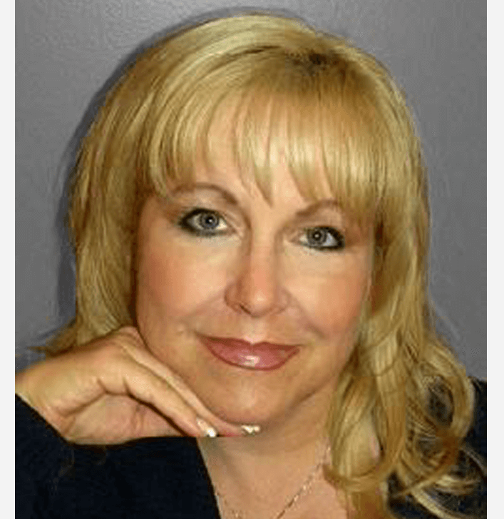 A close-up portrait of a smiling blonde woman resting her chin on her hand against a plain background.