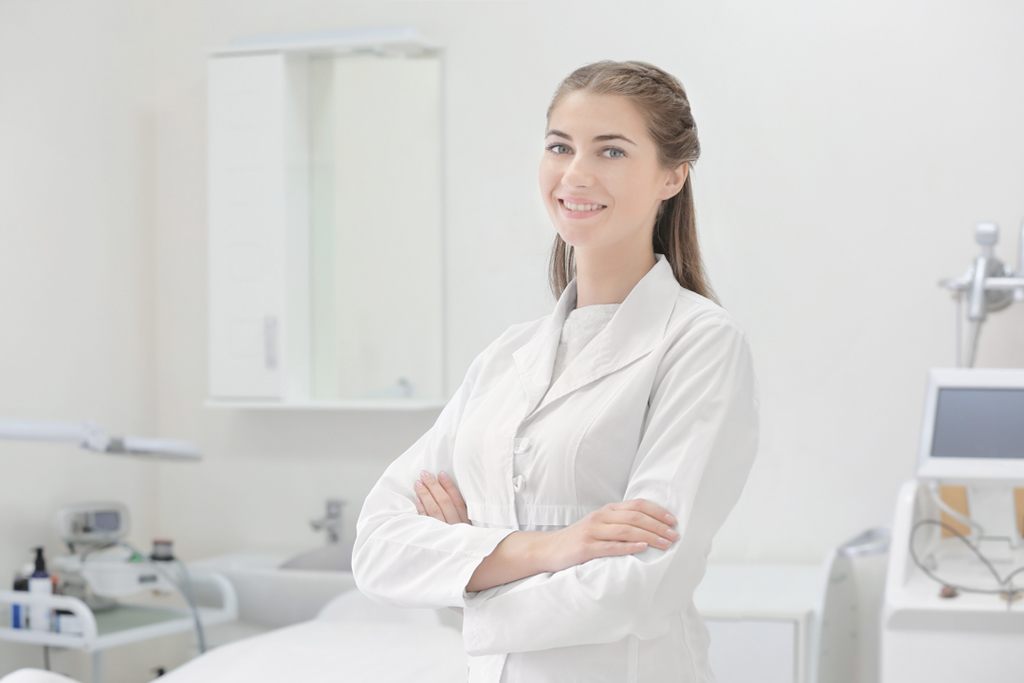 A smiling female medical professional in a white coat standing confidently in a bright clinical setting.