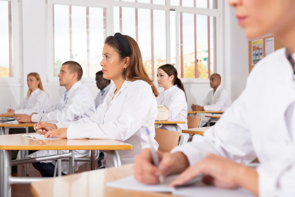 Students in white lab coats attentively listening in a classroom setting, likely during a science or medical lecture.
