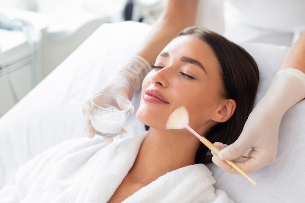 A woman receiving a gentle facial makeup application while lying down, wearing a white robe.